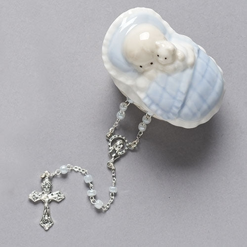A porcelain box in the shape of a baby boy tucked in his crib, with a blue rosette design and a cross, enclosed in a glass box with a silver chain.