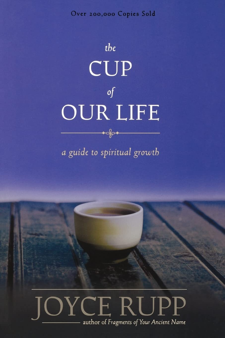 Book cover of 'The Cup of Our Life' by Joyce Rupp with a cup on a wooden surface.