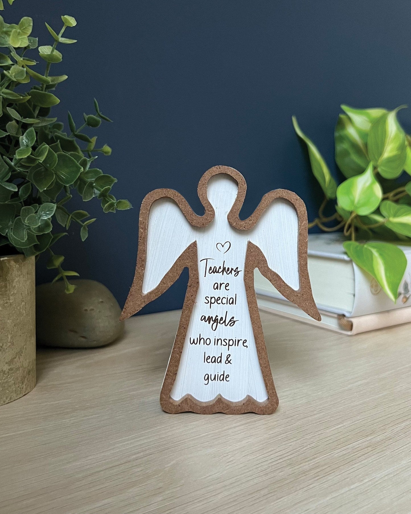 A wooden angel plaque with a message for teachers, displayed on a flat surface with a plant and a book in the background.