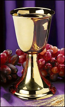 A gold-plated brass communion cup with a stem, displayed in front of a purple background with red grapes scattered around.