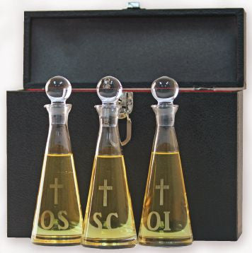 Three clear glass decanters with golden-colored oil inside, placed in a black case with a cross engraving on the side.