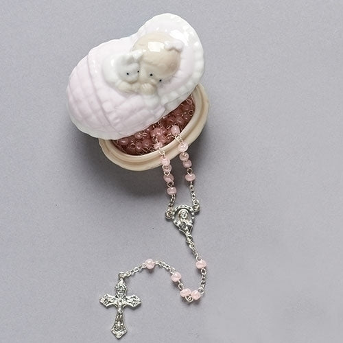 A porcelain box in the shape of a baby girl crib with a pink glass rosary placed beside it.
