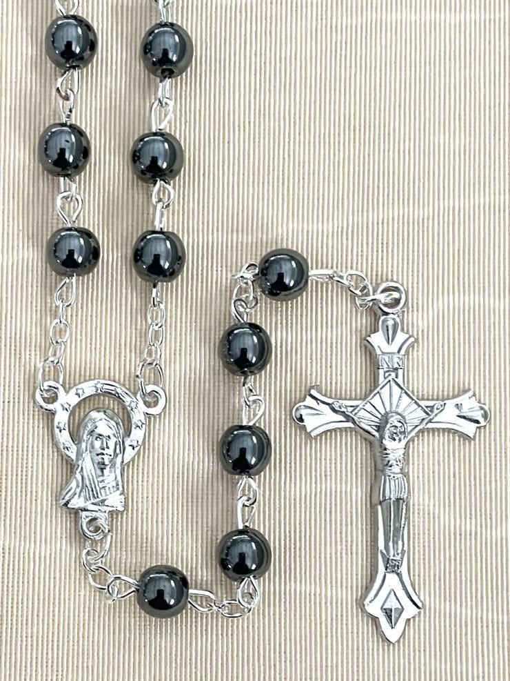 A black and silver rosary necklace featuring round black beads, a silver Miraculous Medal centerpiece, and a detailed silver crucifix, displayed on a textured beige fabric background.