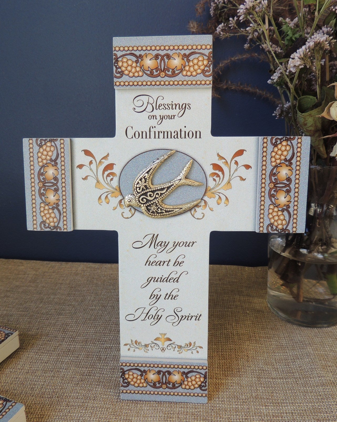 Decorative cross with religious text and design on a table with a vase of flowers in the background.
