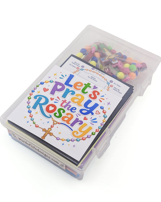 A clear plastic storage box containing a printed card set titled "Let's Pray the Rosary" with colorful beads and small rosary components visible inside, photographed on a white background.