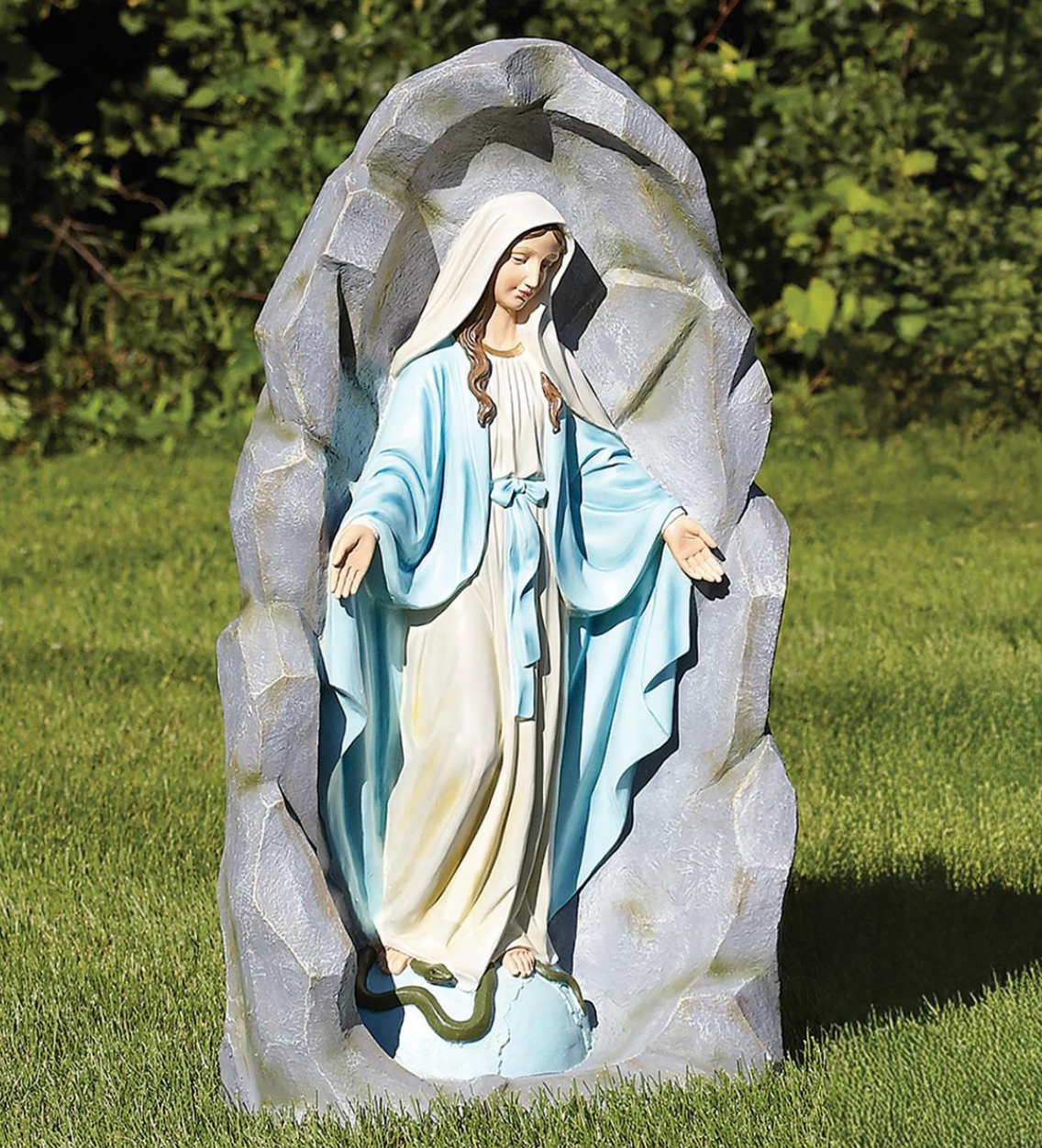 A Mary garden statue standing in front of a faux rock, dressed in a light blue robe and cream gown, placed on a green lawn with foliage in the background.