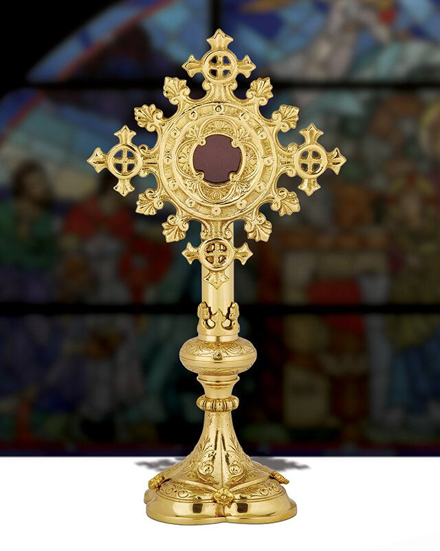A brass reliquary with ornate designs, featuring a 3-inch opening at the top for displaying relics, placed against a blurred background with a religious painting.