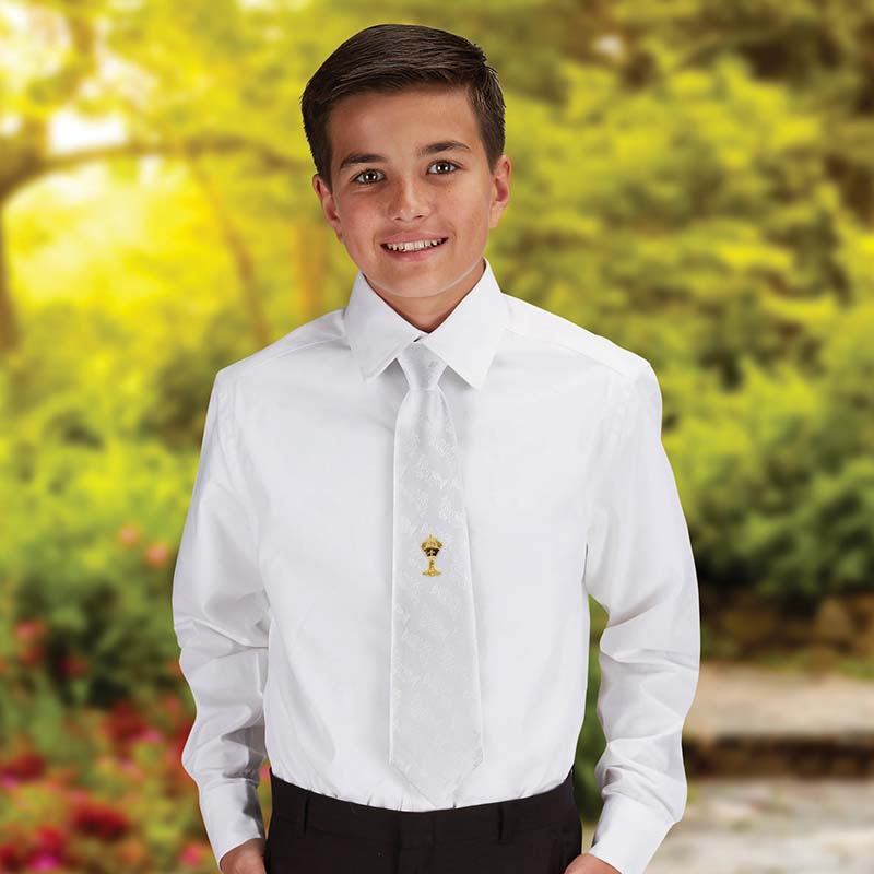 A young boy wearing a white satin brocade tie with a golden chalice design in the middle, standing outdoors with flowers in the background.