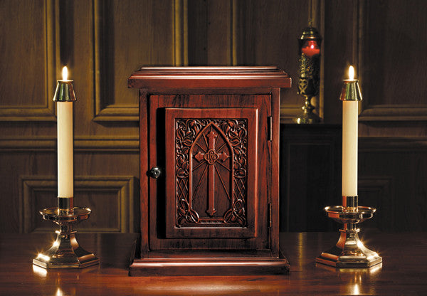 A wooden tabernacle with a carved door and candles on either side, placed on a wooden table with a dark background.