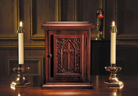 A wooden tabernacle with a carved door and candles on either side, placed on a wooden table with a dark background.