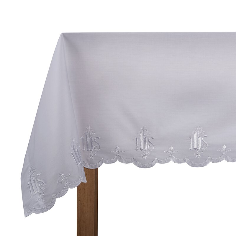 A white altar frontal with embroidered IHS design on a wooden table.