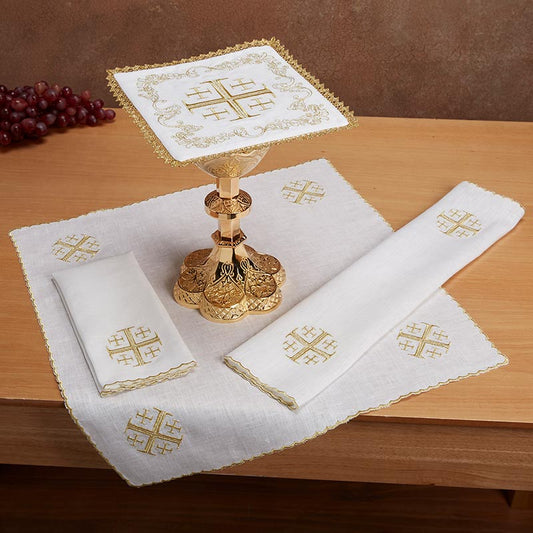 A set of white linen altar cloths with gold embroidered designs, including a chalice pall and a lavabo towel, displayed on a wooden table with a cross design pattern.
