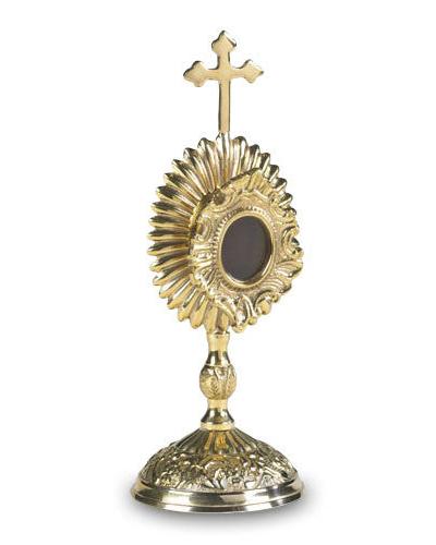 An oval-shaped brass reliquary with a cross on top and a round window in the center, featuring intricate designs on the base and around the central window.