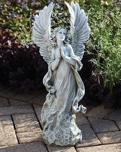 A gray, stone-finished garden statue of a praying angel with wings, placed on a brick pathway with plants in the background.