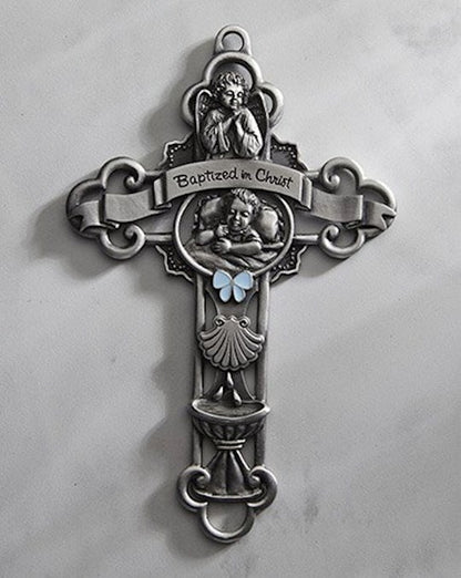 A silver-colored baptism cross with a blue enamelled bow in the centre and the words 'Baptized in Christ' inscribed on it. The cross has intricate hand-sculpted details.